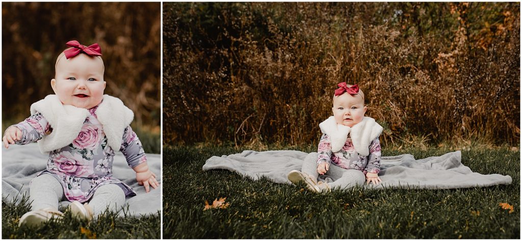 A fall family photo session featuring a six-month-old girl outside her home in New Jersey. She is sitting up unsupported on a grey blanket outside. The fall foliage is vibrant behind her. She is wearing a faux fur jacket, floral dress, and a red hair bow.