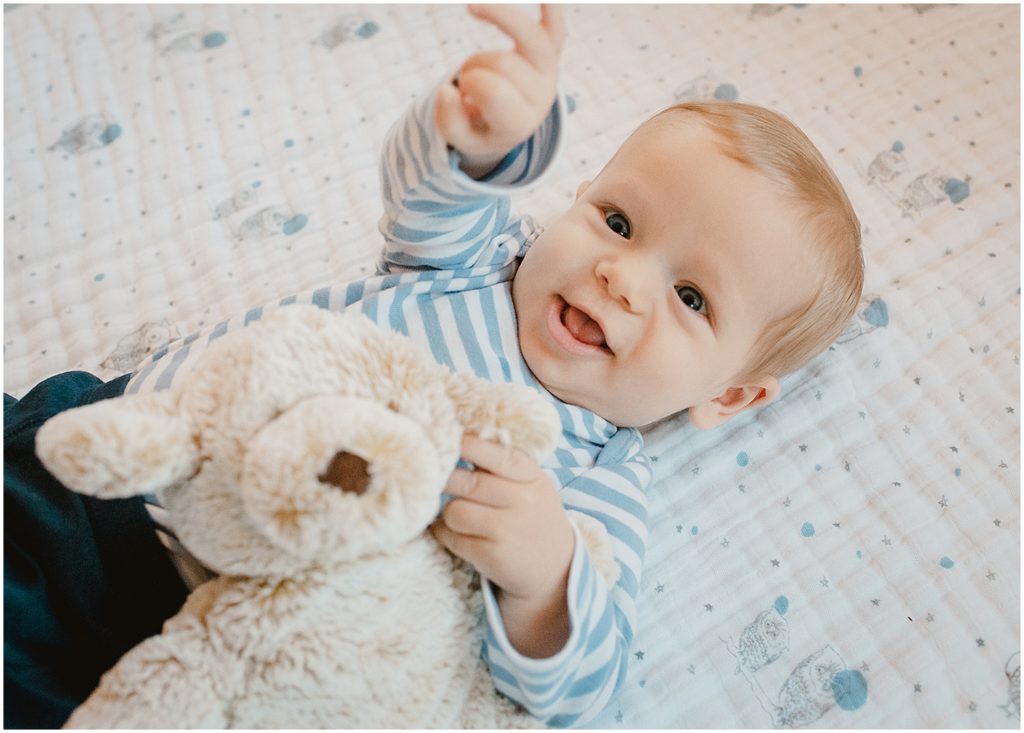 An overhead photo of a six-month-old boy at a lifestyle family session in a Brooklyn brownstone apartment. He reaches up toward the camera, smiling, as he holds onto his favorite stuffed animal toy bear.