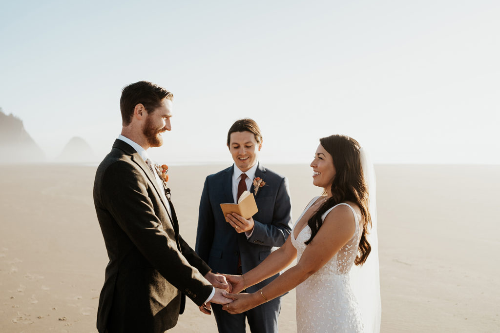 Sam, in a white dress and veil, holds hands with Andrew on an empty beach on the Oregon coast. Sam's brother Matt stands just behind them, officiating their elopement.
